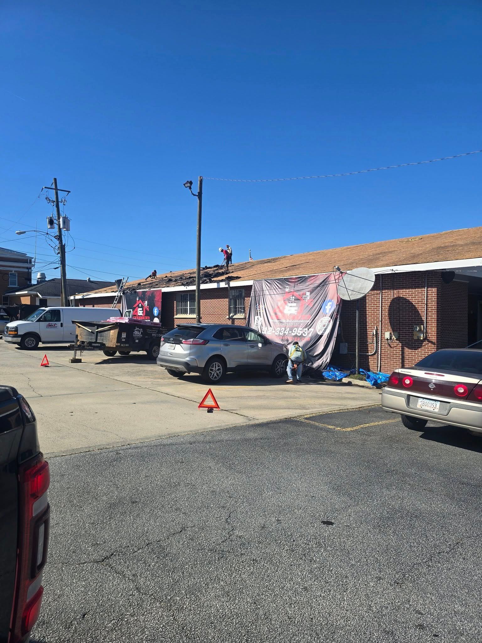 A person works on a building's roof with a banner and vehicles in a parking lot on a sunny day.