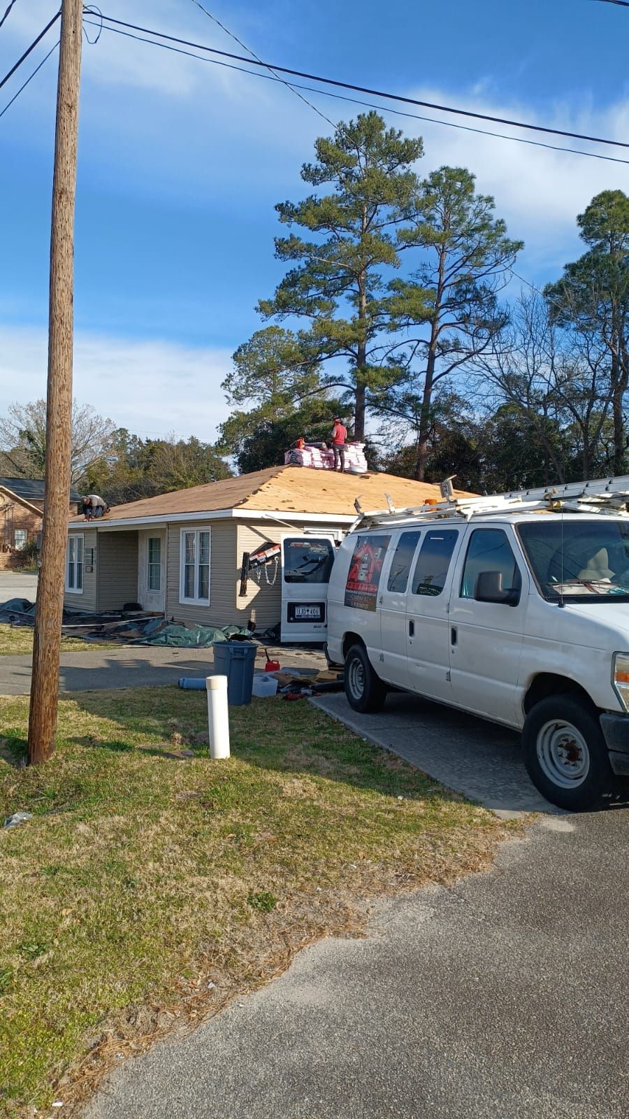 Workers replacing the roof of a one-story house. A white van is parked in the driveway. The sky is blue.