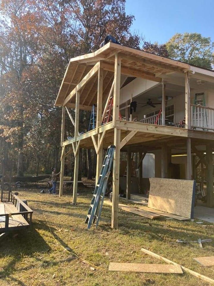 Construction of a two-story wooden house with a porch. Workers are on the roof and ground. The setting is outdoors with trees in the background.