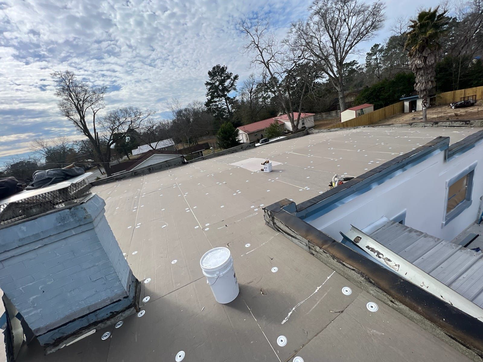 A flat roof with paint spots and a white bucket, overlooking a neighborhood with trees under a cloudy sky.
