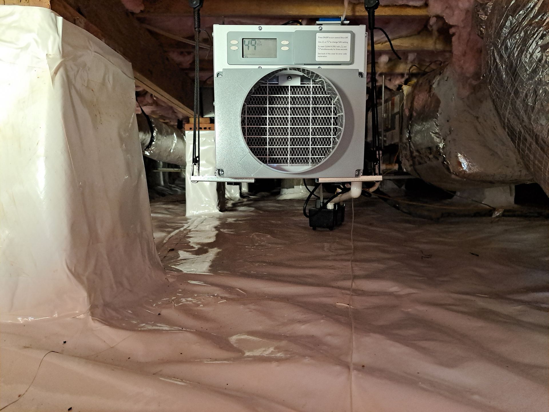Dehumidifier suspended in a crawlspace, resting on white plastic sheeting. Ductwork and insulation visible.