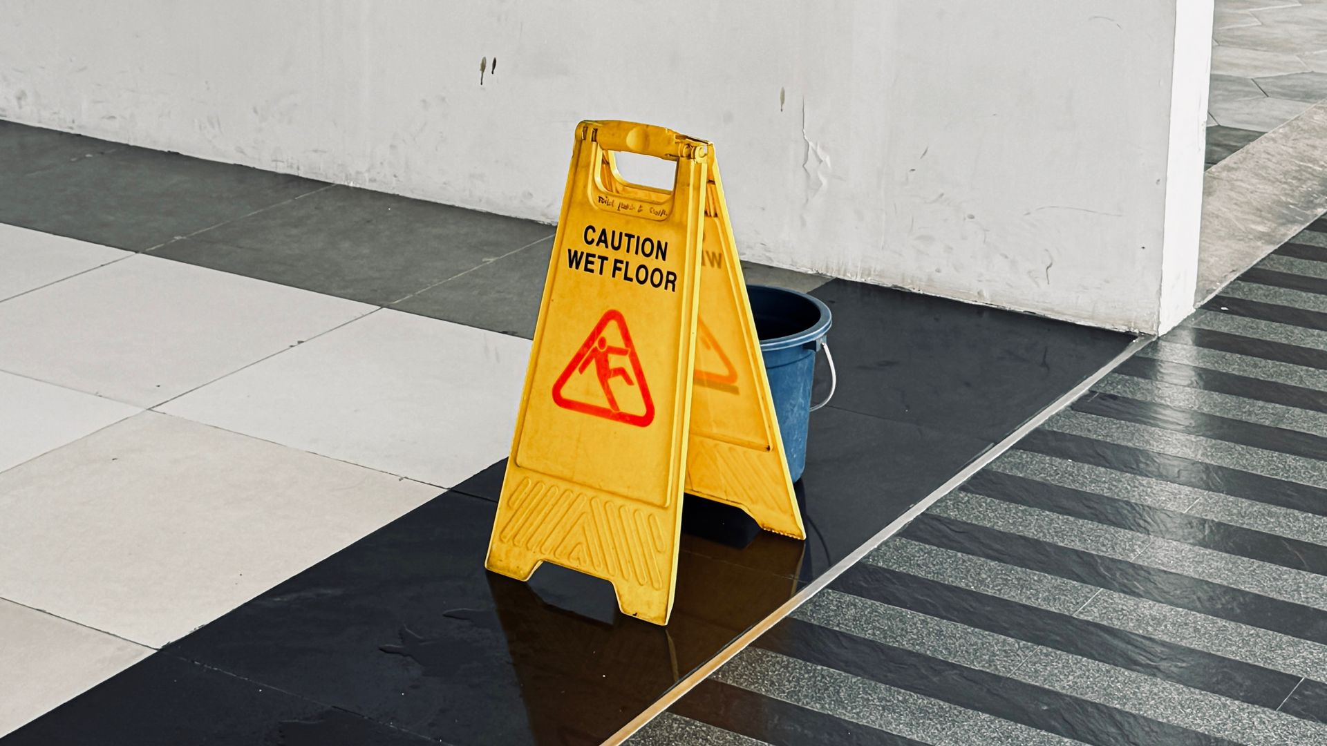 A yellow caution sign is sitting on a wet floor next to a bucket.