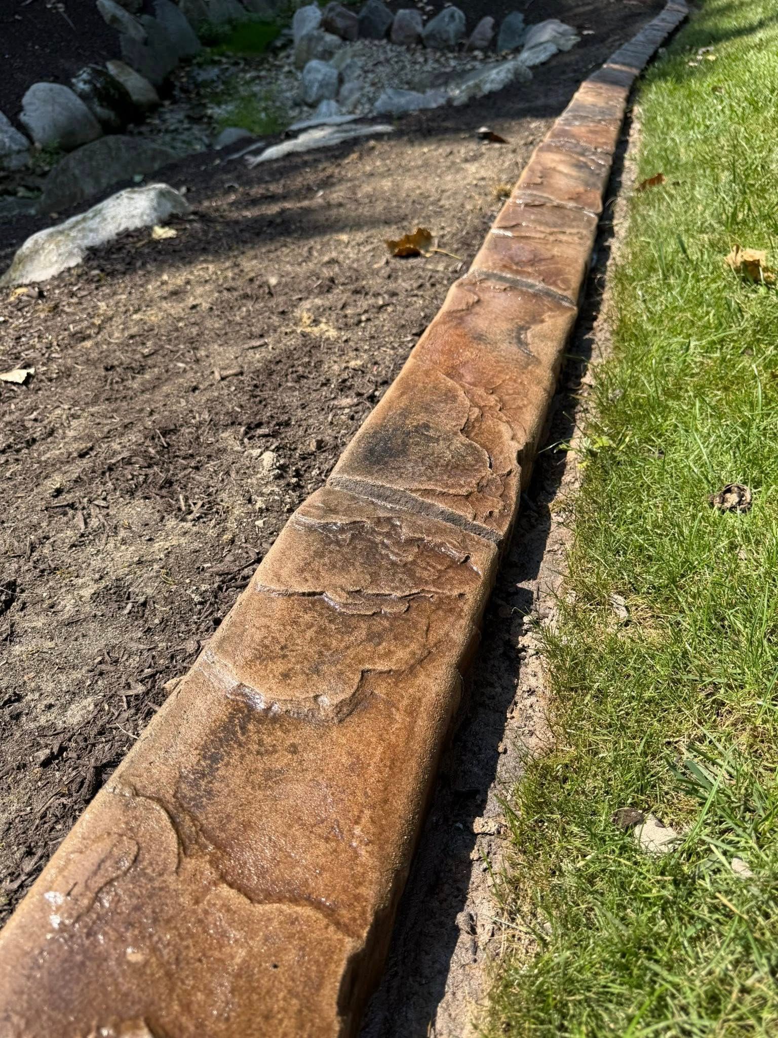 A decorative stamped concrete landscape border separates a patch of dirt and large rocks from a vibrant green lawn.