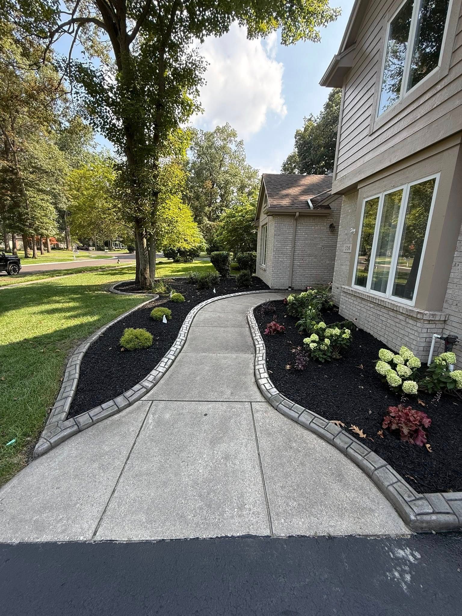 A concrete sidewalk leads to a house entrance, bordered by black mulch, small shrubs, and gray stone edging.