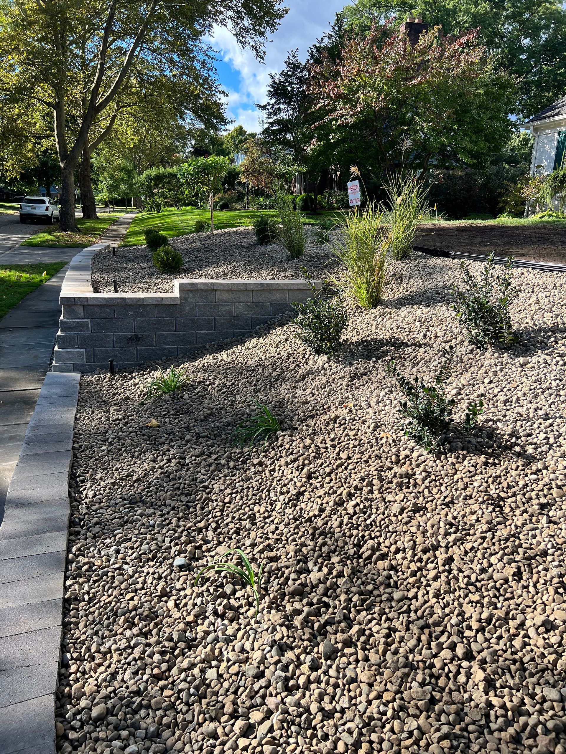 A stone retaining wall tiers a landscaped yard filled with gravel, small shrubs, and mulch under a bright, sunny sky.