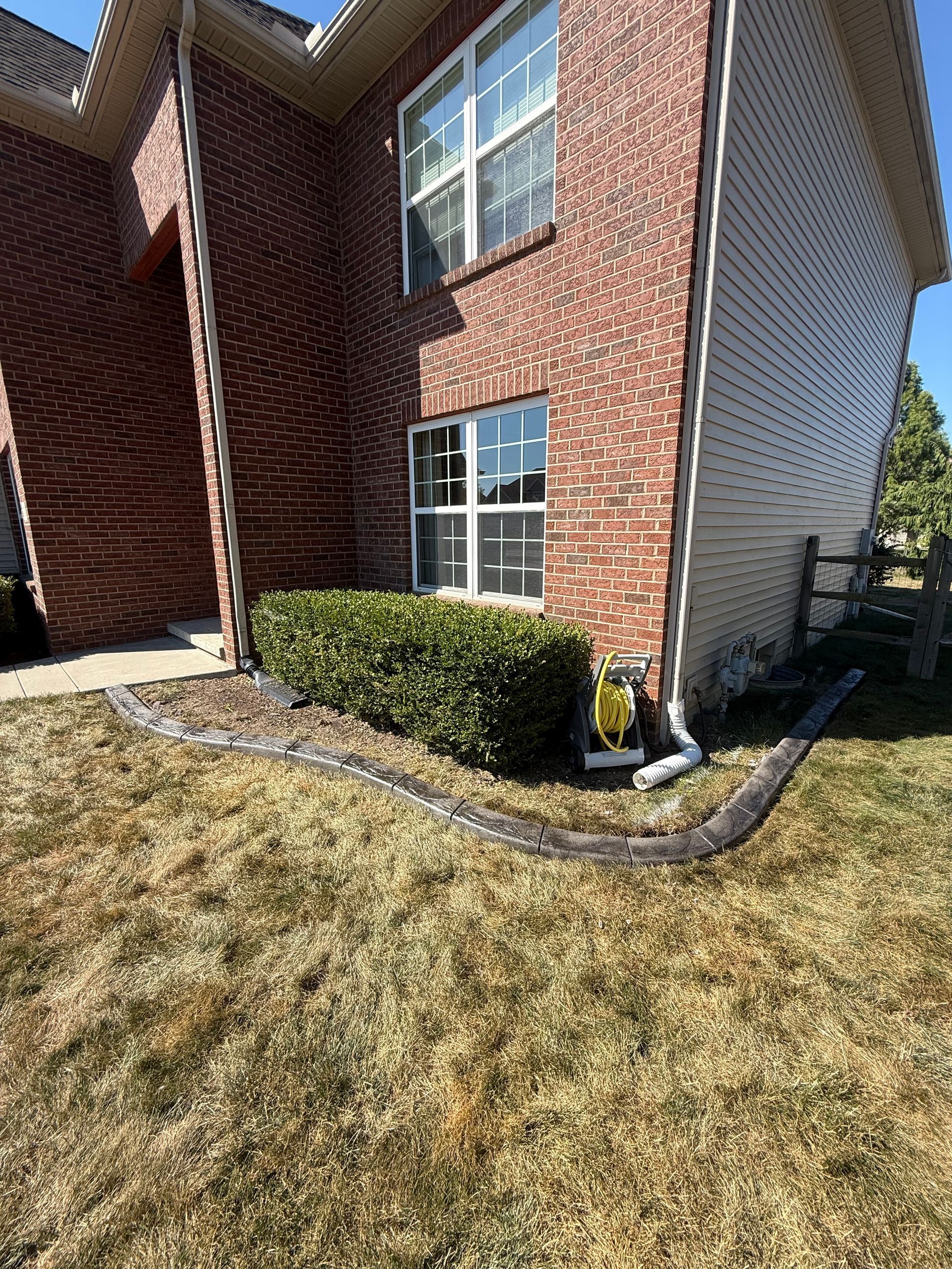 A red brick house exterior features a small green hedge in front, with a black landscape drain pipe resting on the lawn.