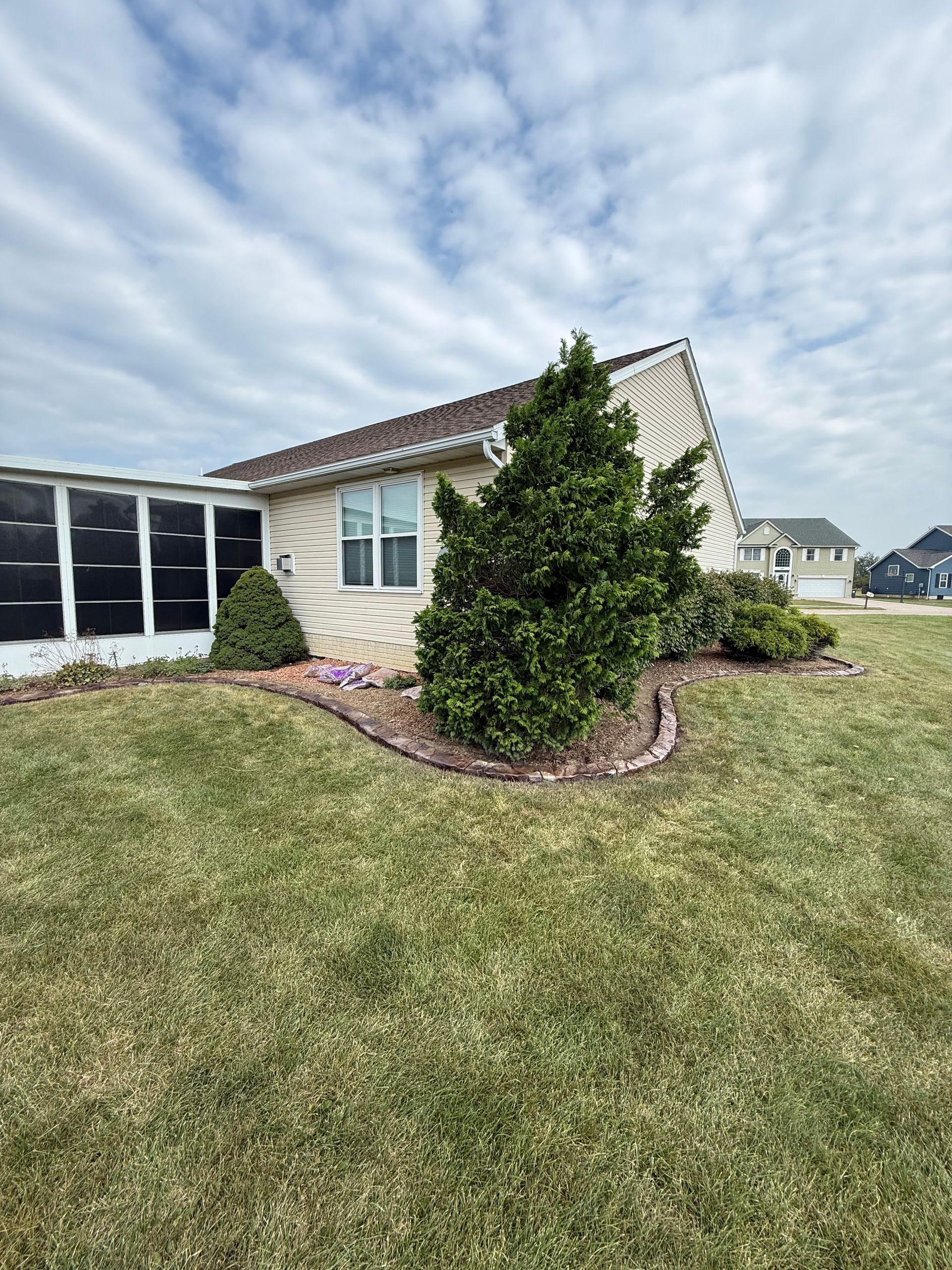 A suburban home featuring light beige siding, a screened-in porch, and a landscaped garden bed with evergreen shrubs.