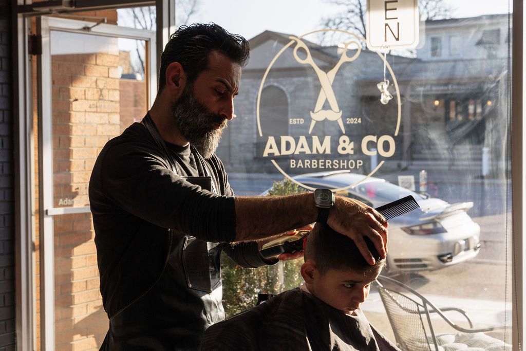 A man is cutting a boy 's hair in a barber shop with a logo on the window.