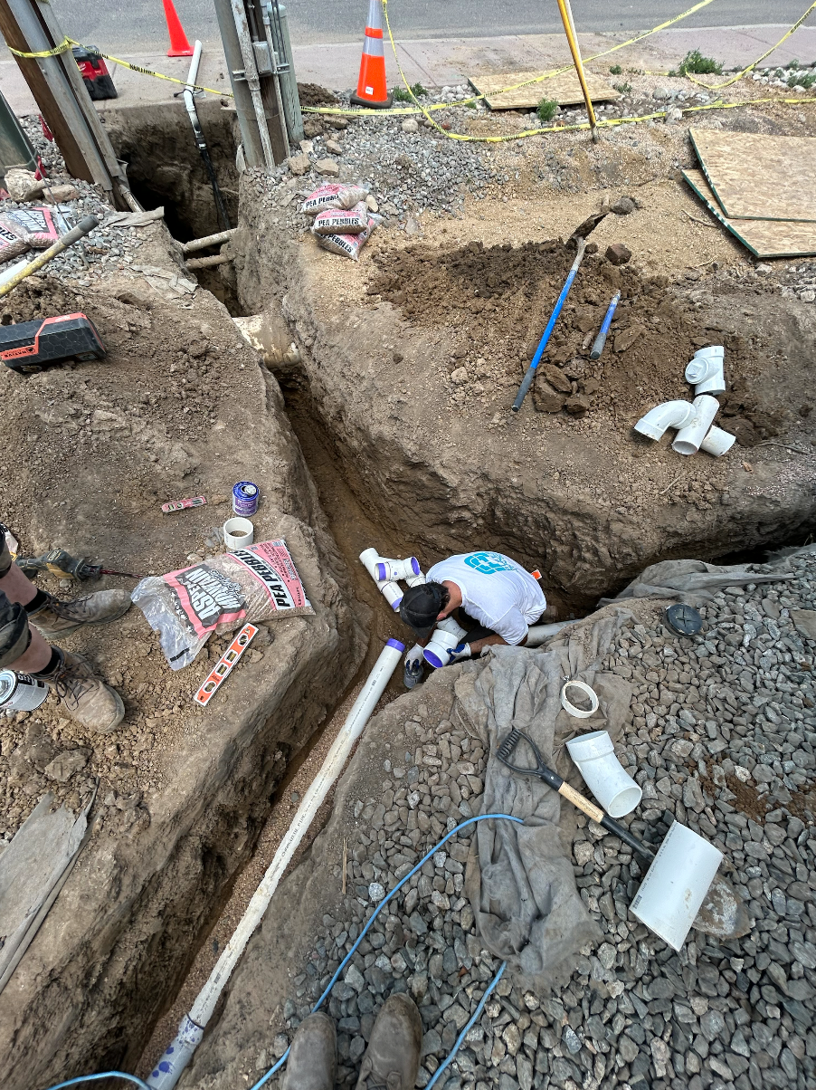 Construction worker in a trench, installing PVC pipes. Dirt, tools, and caution cones visible.