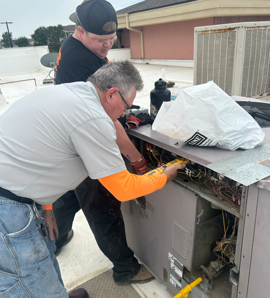 Two HVAC technicians working on rooftop unit. One uses a yellow tool on wiring.