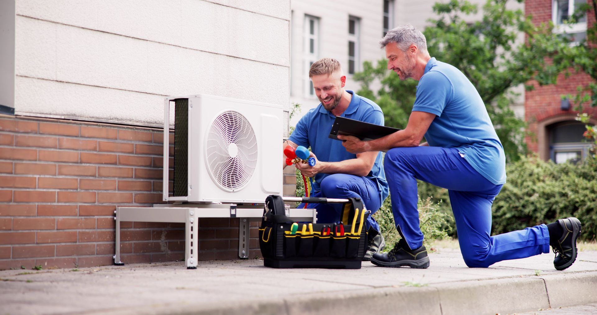 Two technicians in blue uniforms fixing an AC unit, one kneeling with a tablet, the other holding a tool.