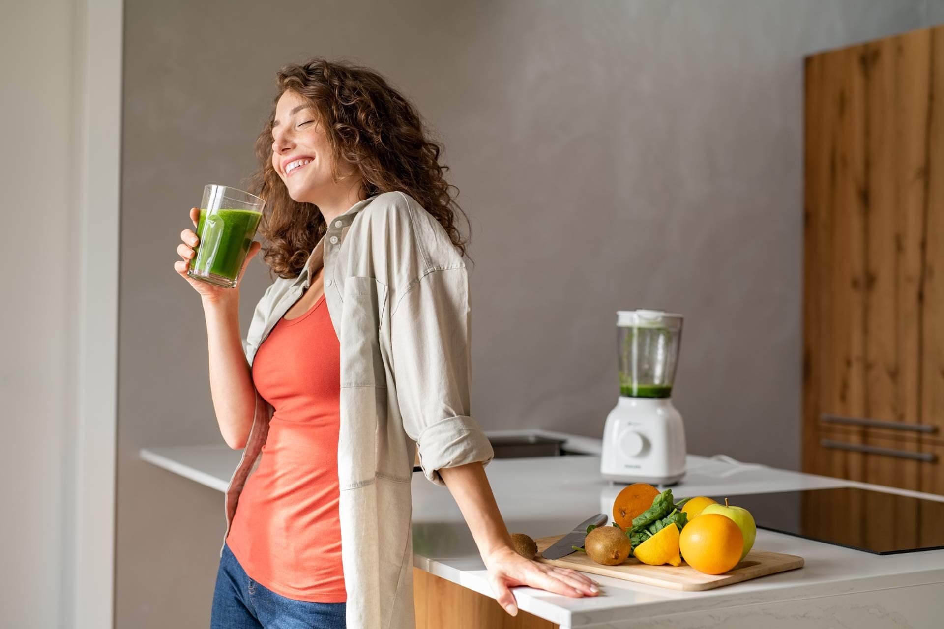 A pregnant woman is drinking a green smoothie in a kitchen.
