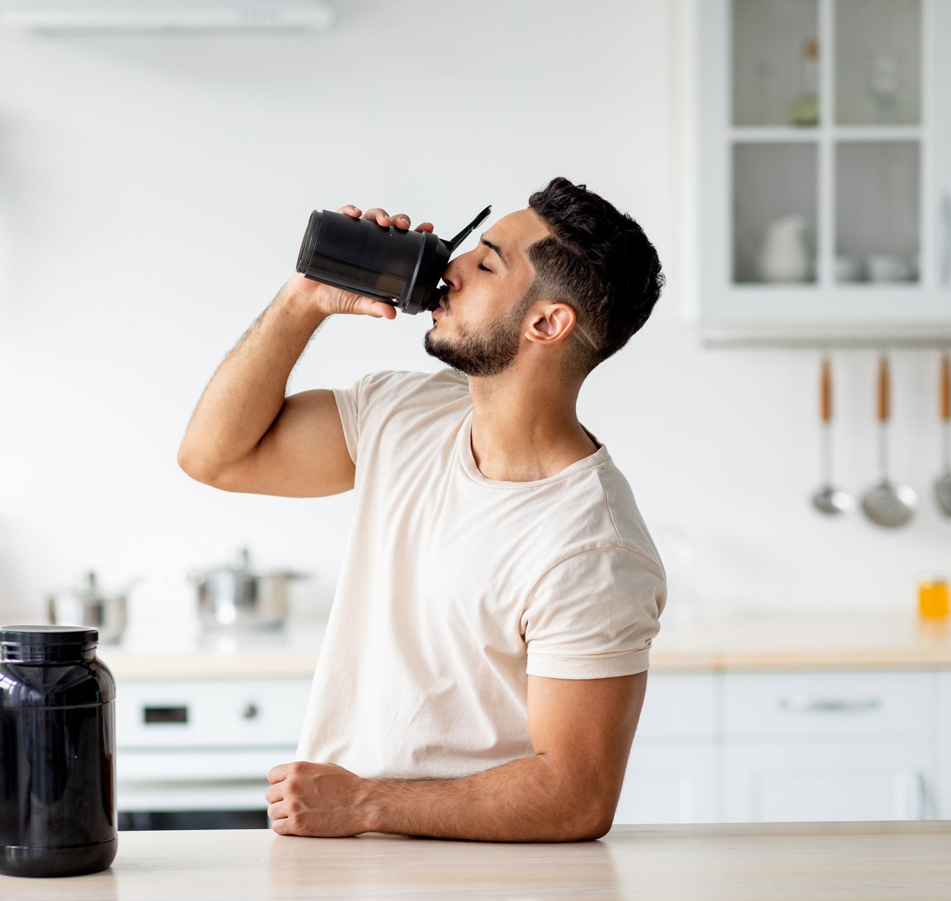 A man is drinking a protein shake from a shaker in a kitchen.