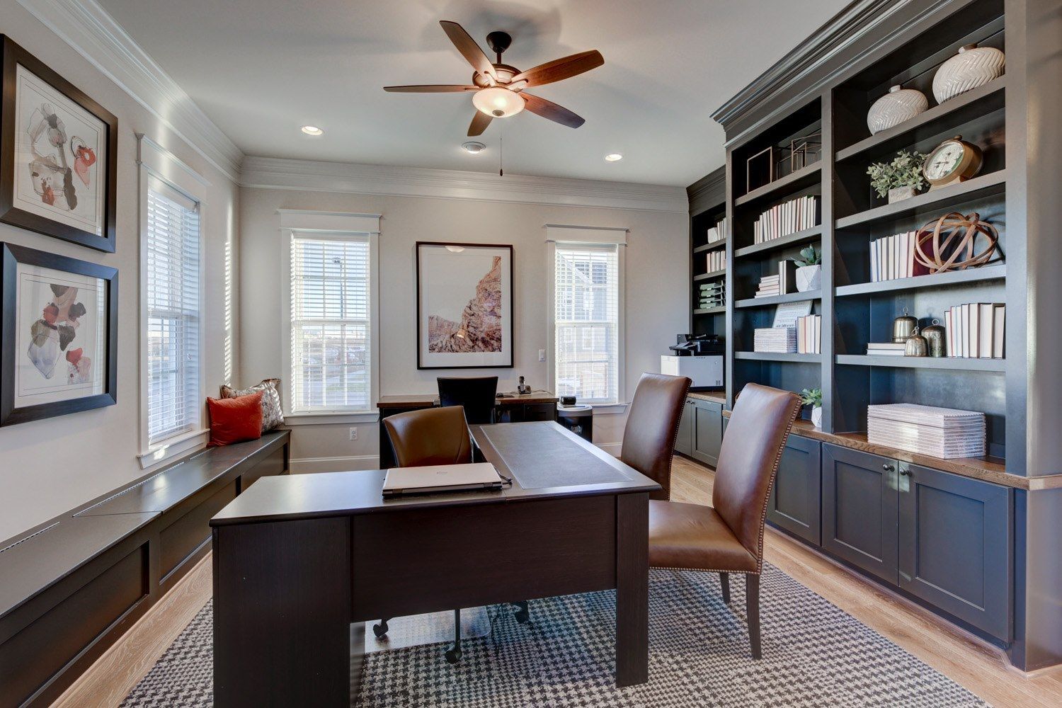 A home office with a desk , chairs , shelves and a ceiling fan.