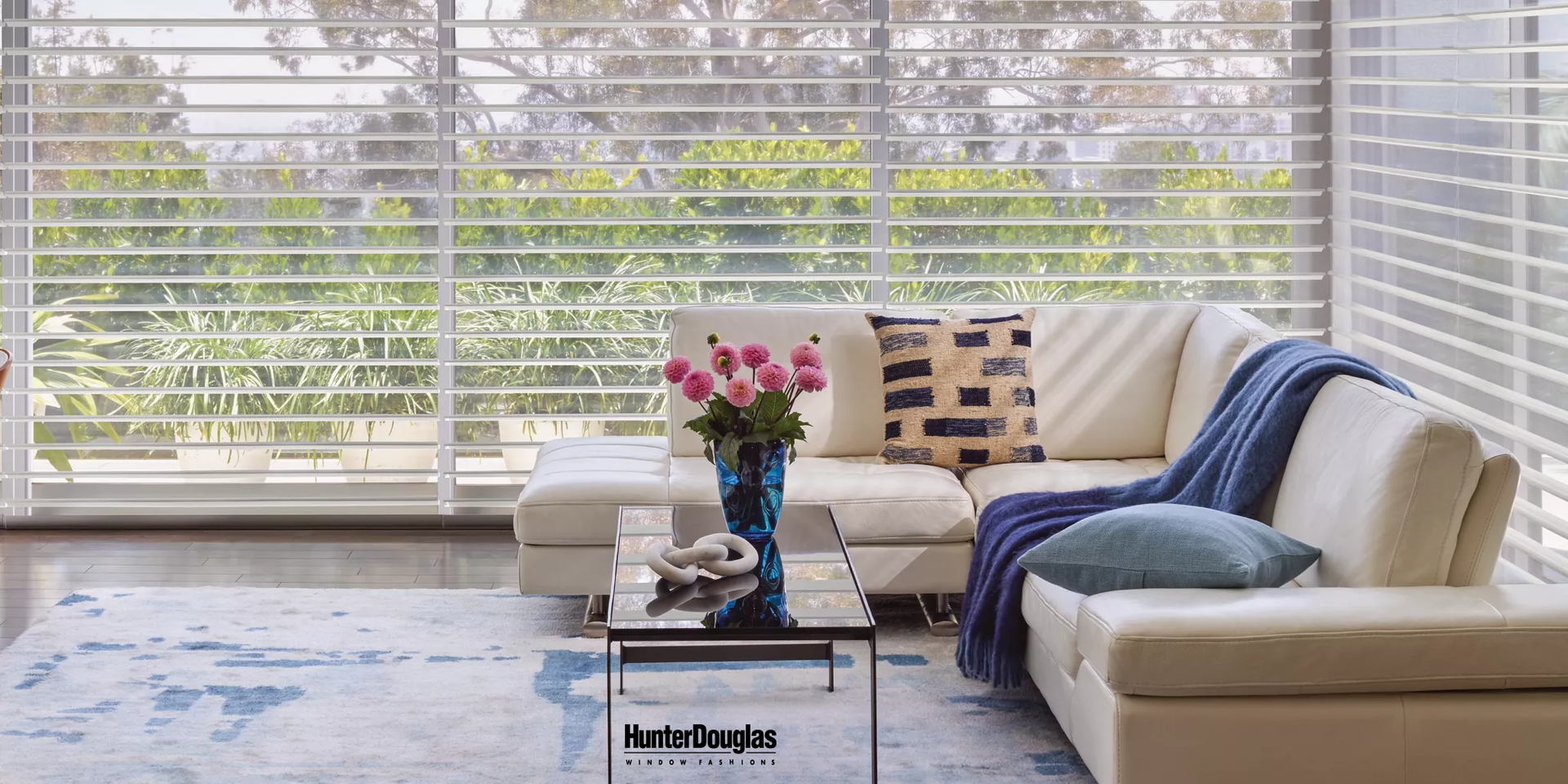 A living room with a couch , coffee table , vase of flowers and blinds.