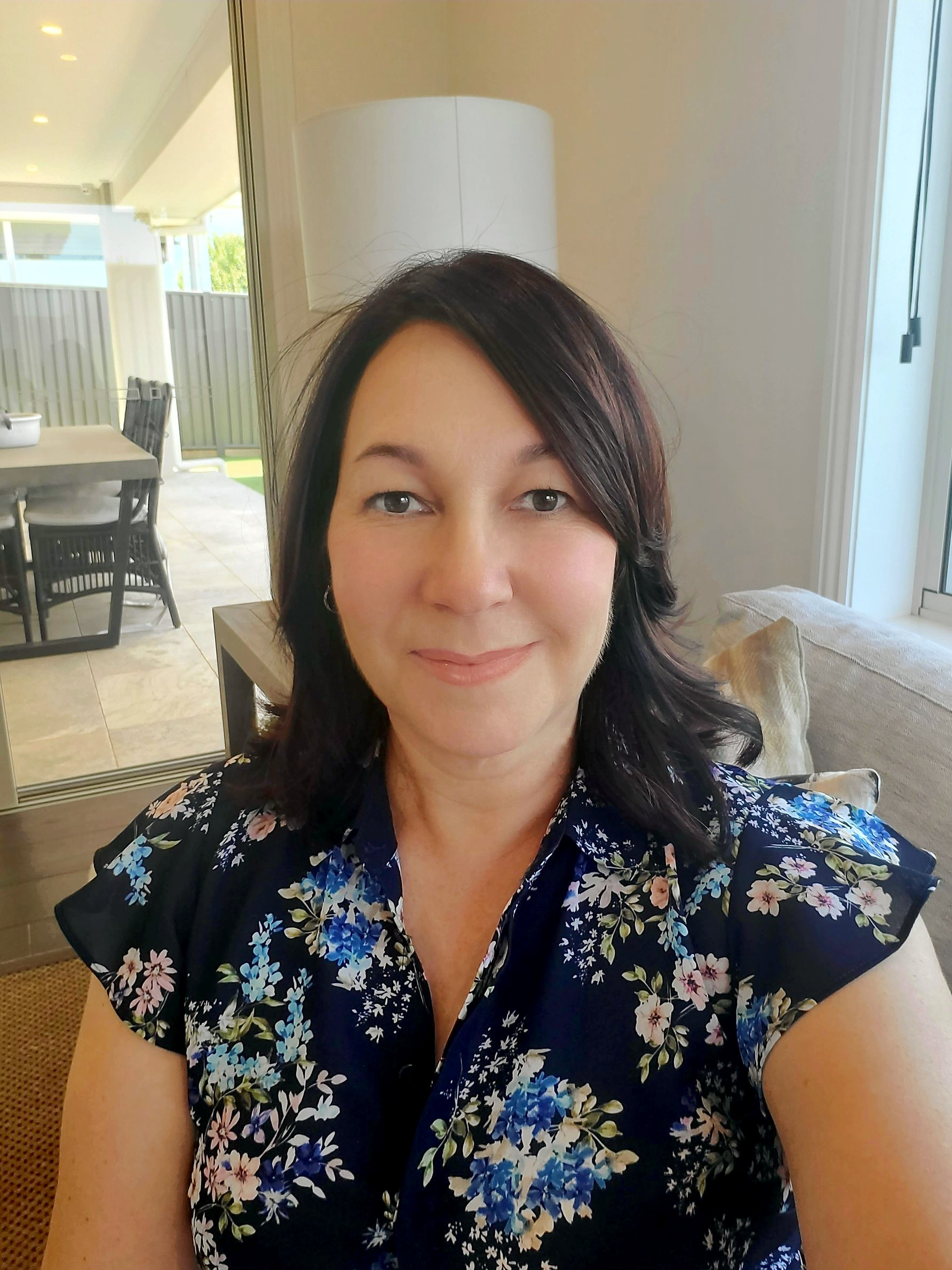 Woman with dark hair in a floral top smiles indoors; a dining area is visible behind her — Jodi Maree Interiors in Dubbo, NSW