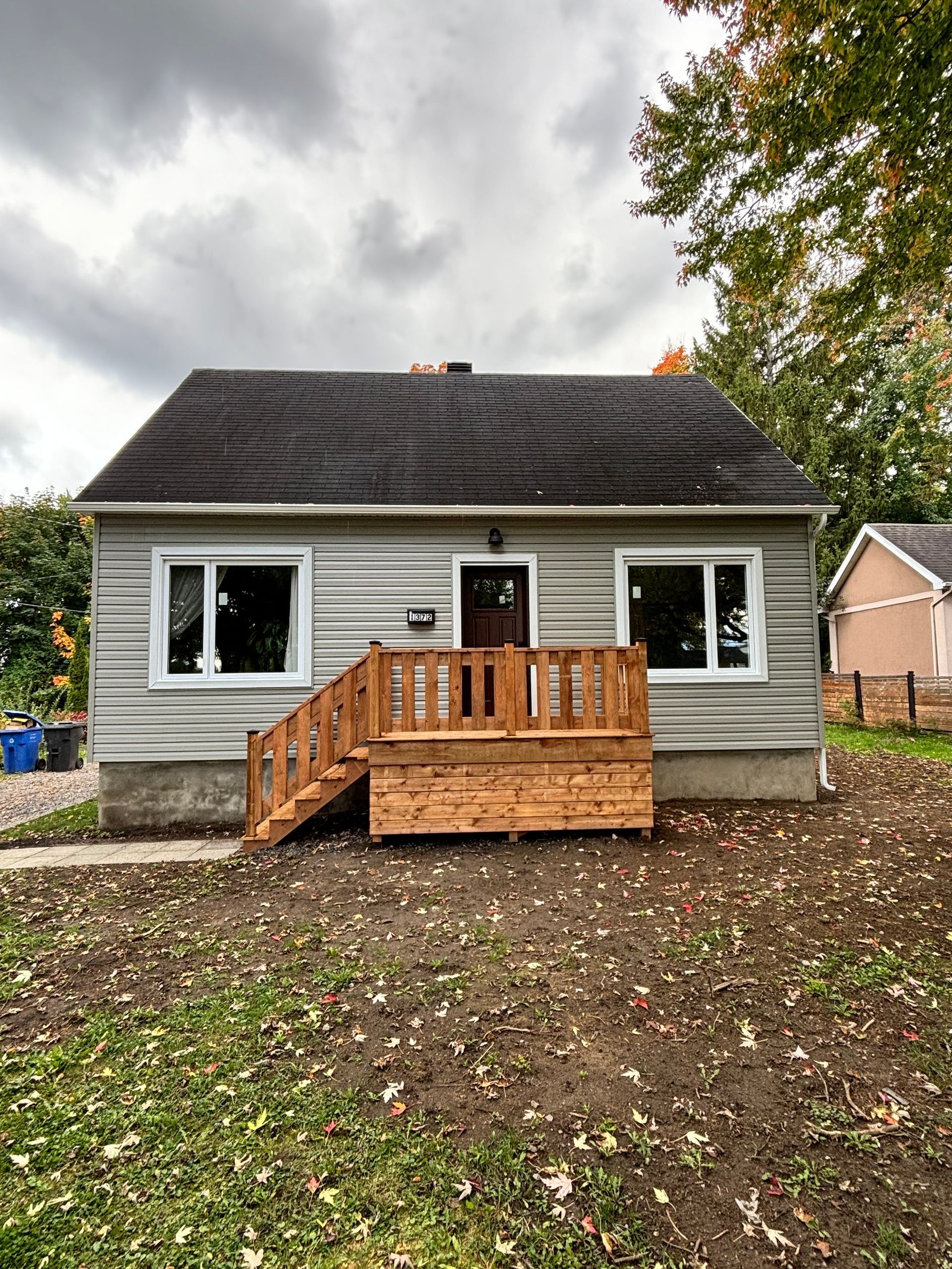 Petite maison grise avec un porche en bois et deux fenêtres, se détachant sur un ciel nuageux. La cour n'est pas pavée.