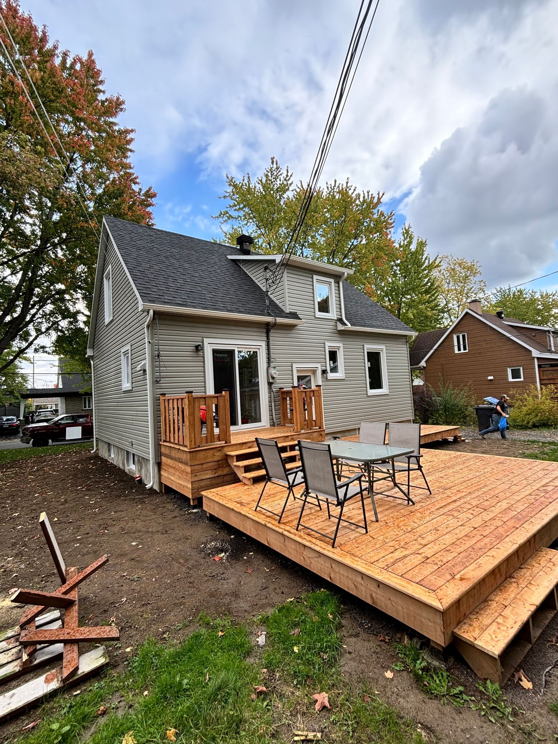 Arrière d'une maison à deux étages avec une terrasse en bois récemment construite. Une table et des chaises se trouvent sur la terrasse inférieure, et un petit jardin s'ouvre devant.