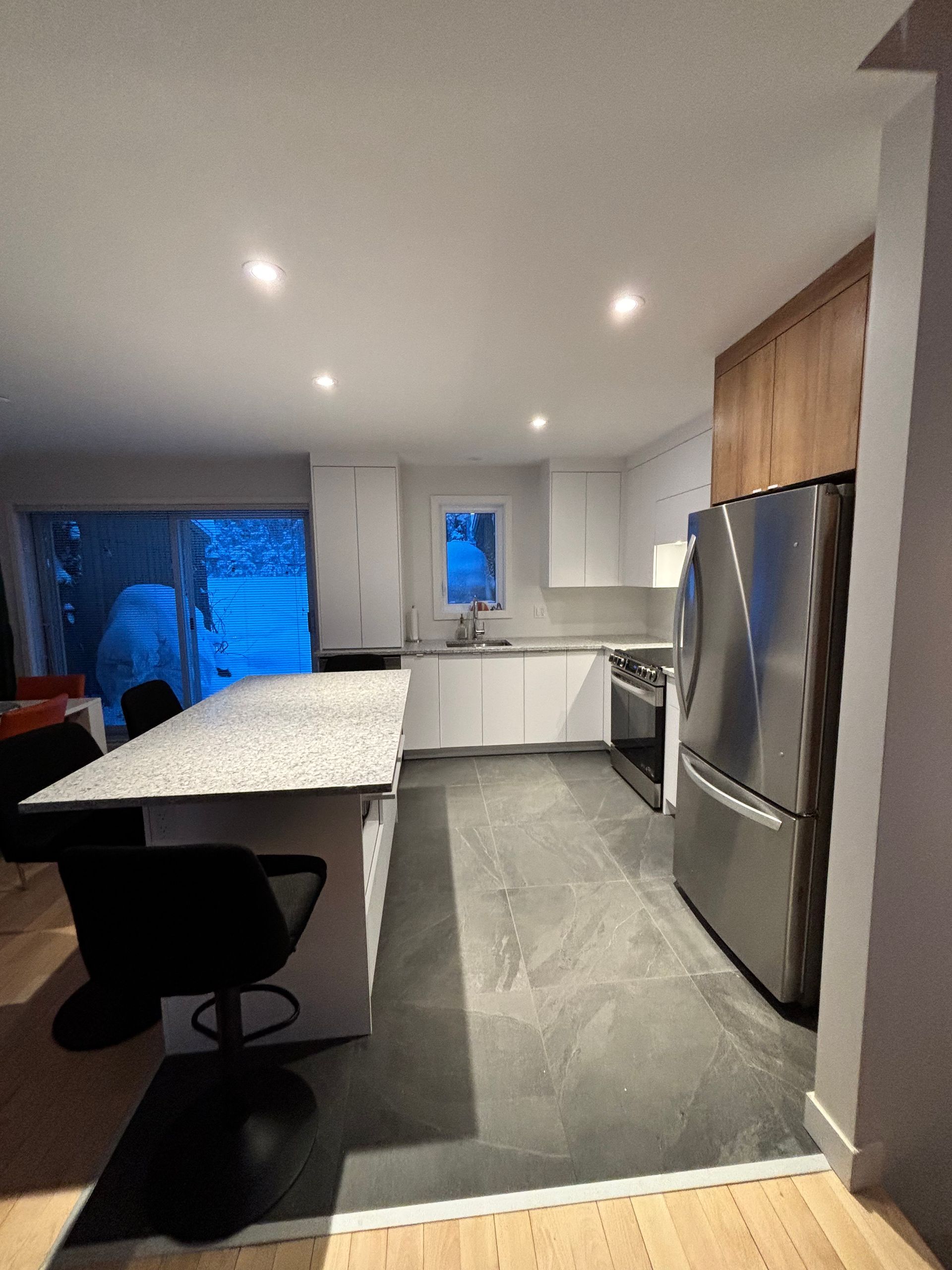 Cuisine moderne avec armoires blanches, îlot central avec comptoir en pierre et tabourets de bar noirs. Électroménagers en acier inoxydable et carrelage foncé.