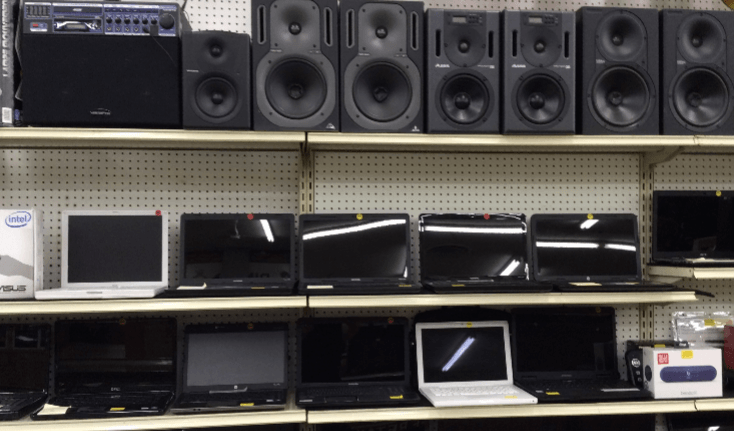 Shelves stocked with speakers, and laptops of varying sizes, against a white wall, possibly in a store.