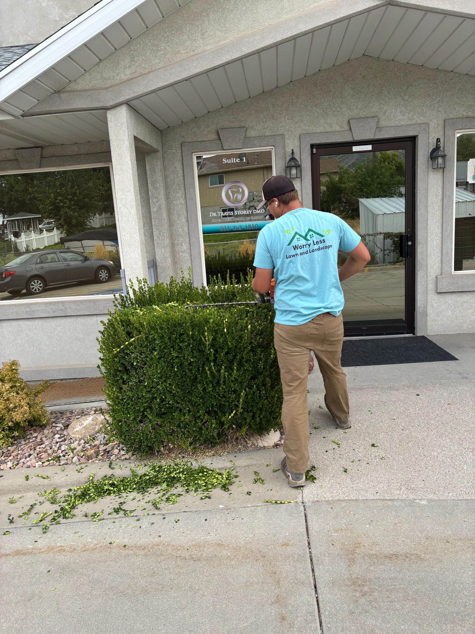 Man trimming a bush outside a building with a black door, wearing a blue shirt and khaki pants.