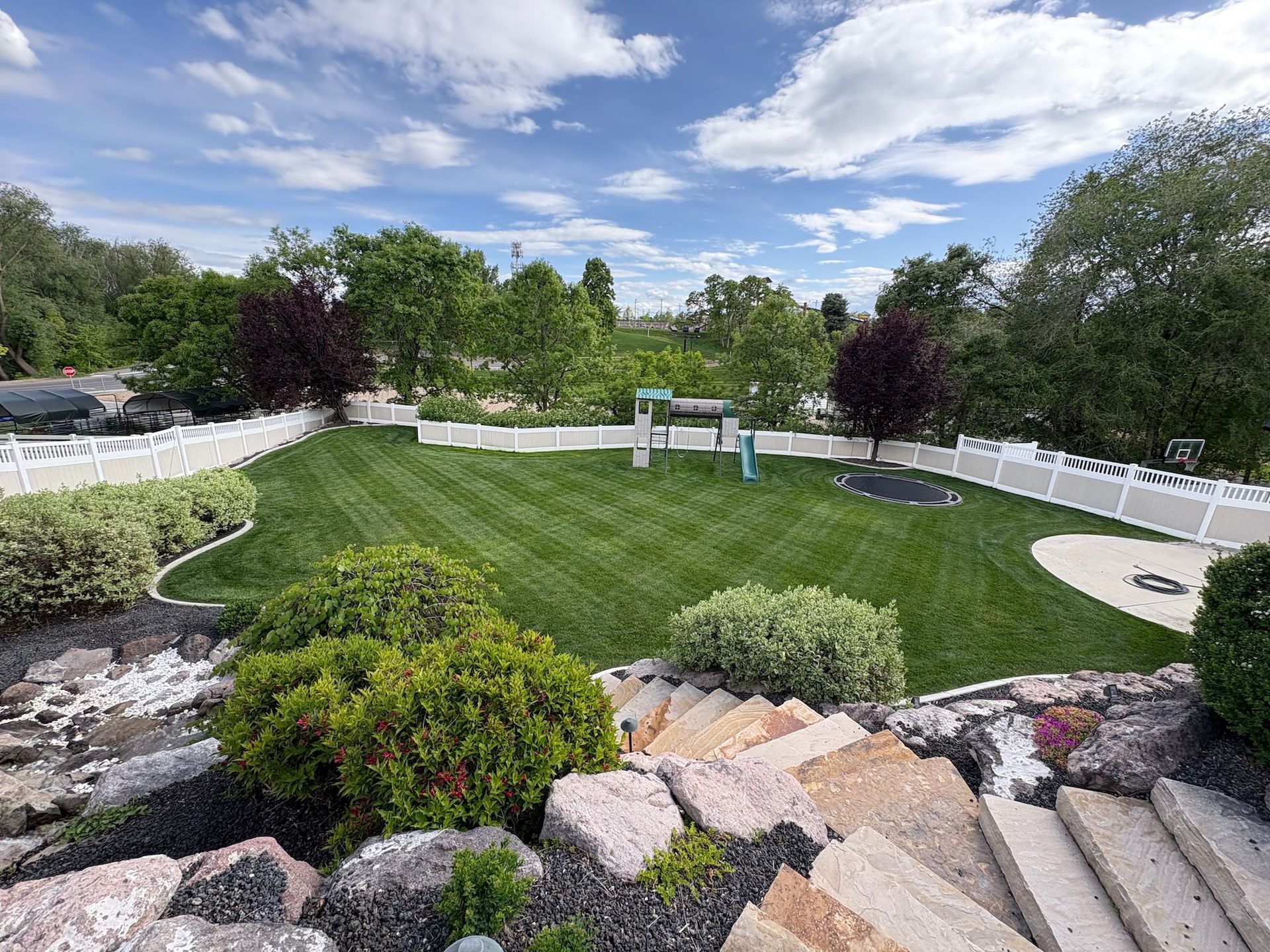 Lush backyard with green lawn, white fence, and a playground under a partly cloudy blue sky.