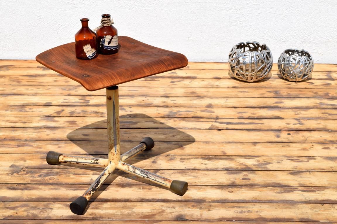 Antiker Arzthocker mit starker Patina am Metallgestell und ergonomisch geformter Sperrholz-Sitzfläche, präsentiert mit historischen Apothekerflaschen auf rustikalem Dielenboden.