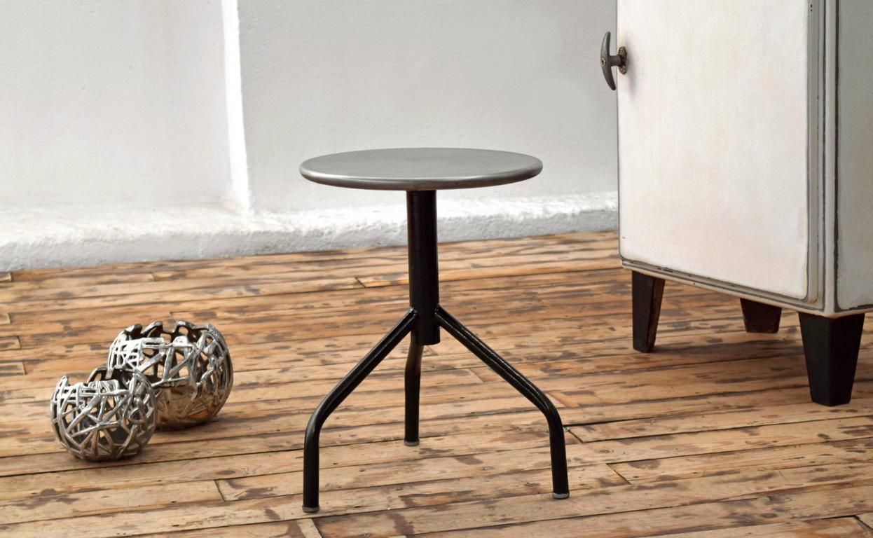 Schwarzer Arzthocker aus Stahlrohr mit runder Metall-Sitzfläche, präsentiert in hellem Interieur auf rustikalem Dielenboden neben einem historischen Medizinschrank