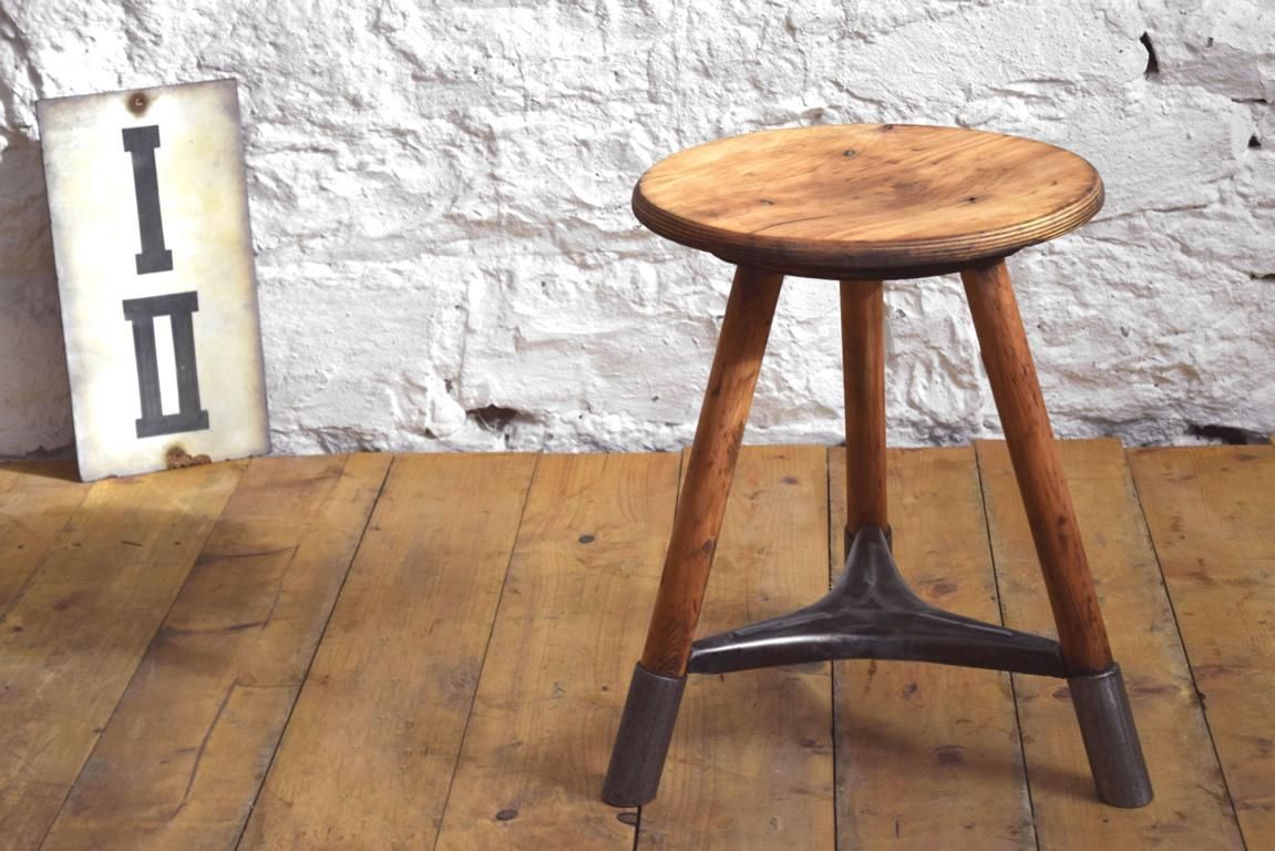 Antiker Dreibeinhocker, mitteldeutsches Industriedesign, Kombination aus Massivholzbeinen und gusseisernem Fußkreuz vor weißer wand
