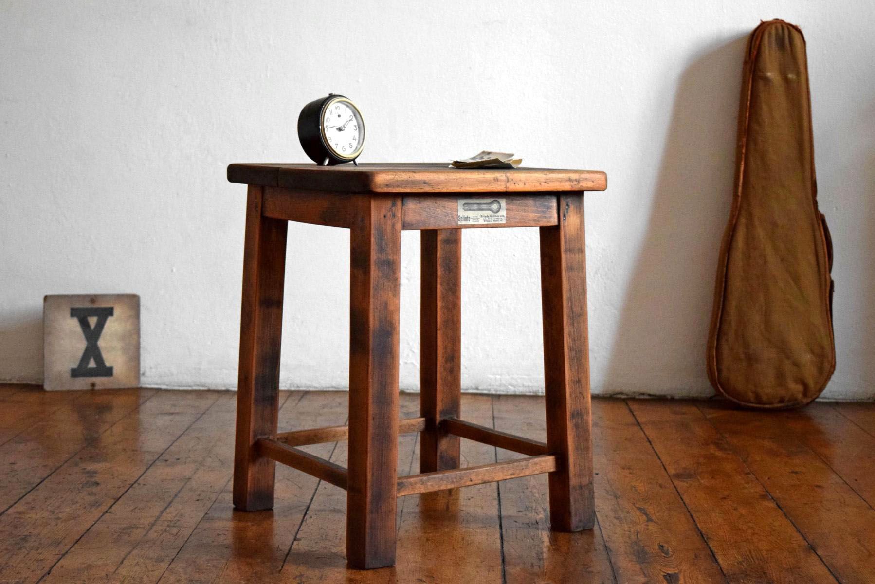Massiver hölzerner Werkstatthocker mit historischer Markung in warmem Wohnambiente, Fokus auf patinierte Holzoberfläche neben einer Geigentasche und einem antiken Wecker