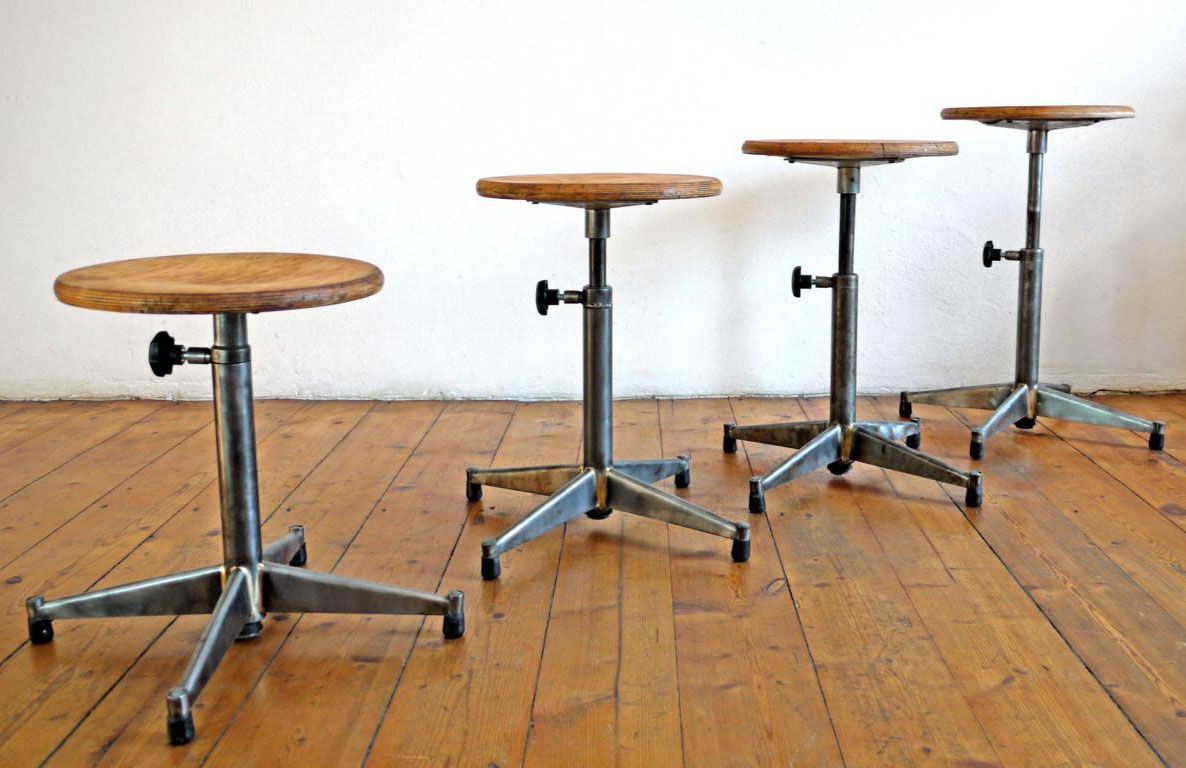 Vier höhenverstellbare Werkstatthocker der späten Moderne auf Holzdielen, Stahlrohr-Konstruktion mit Bakelit-Details und Holzsitzflächen, Beispiel für zeitloses Industriedesign