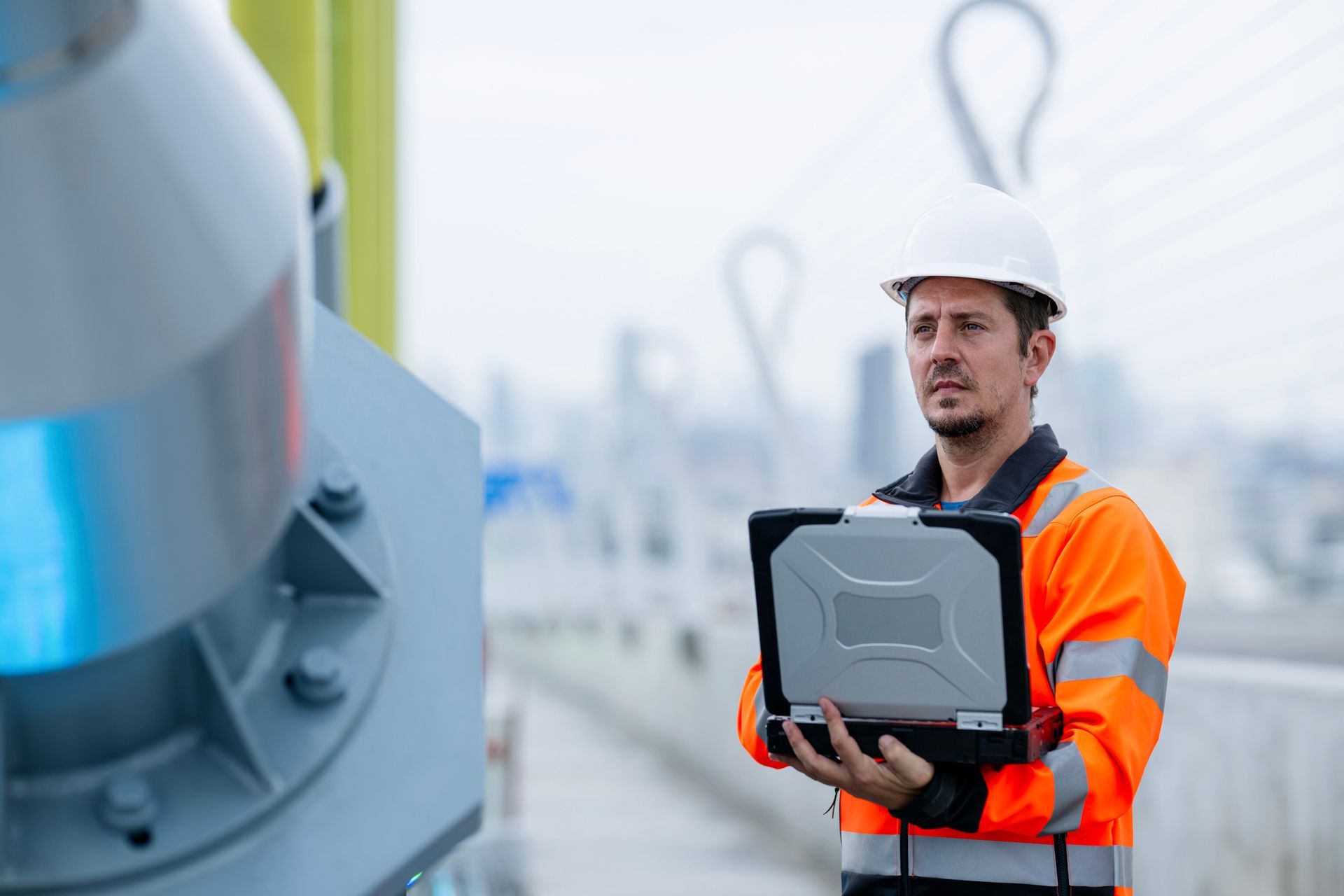Construction worker in hard hat and orange vest holding a laptop, inspecting a structure outdoors.