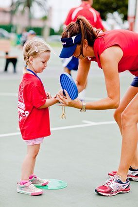 A woman is helping a little girl with her shoes on a tennis court.