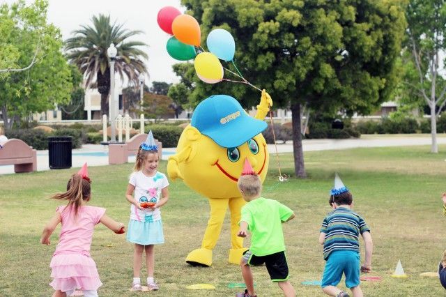 A group of children are playing in a park with a mascot holding balloons.