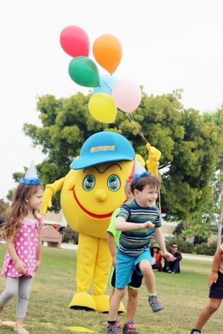 A group of children are playing with a yellow mascot holding balloons.