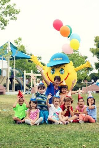A group of children are sitting in the grass with a mascot holding balloons.