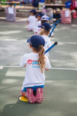 A group of children are kneeling down on a tennis court holding bats.
