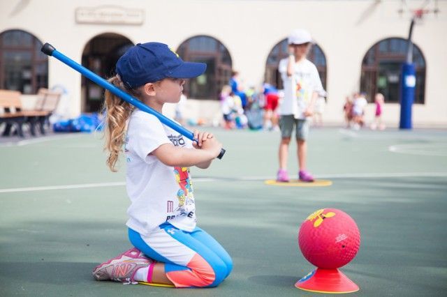 A little girl is kneeling down on the ground holding a bat and a ball.