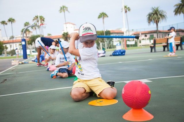 A young boy is kneeling on a court with a ball and a stick.