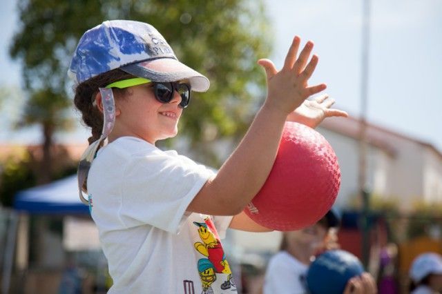A young girl wearing sunglasses and a hat is throwing a red ball.