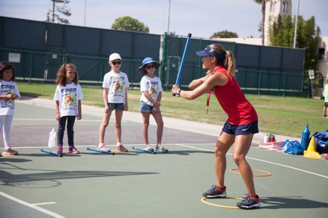 A woman is teaching a group of young girls how to swing a bat on a tennis court.