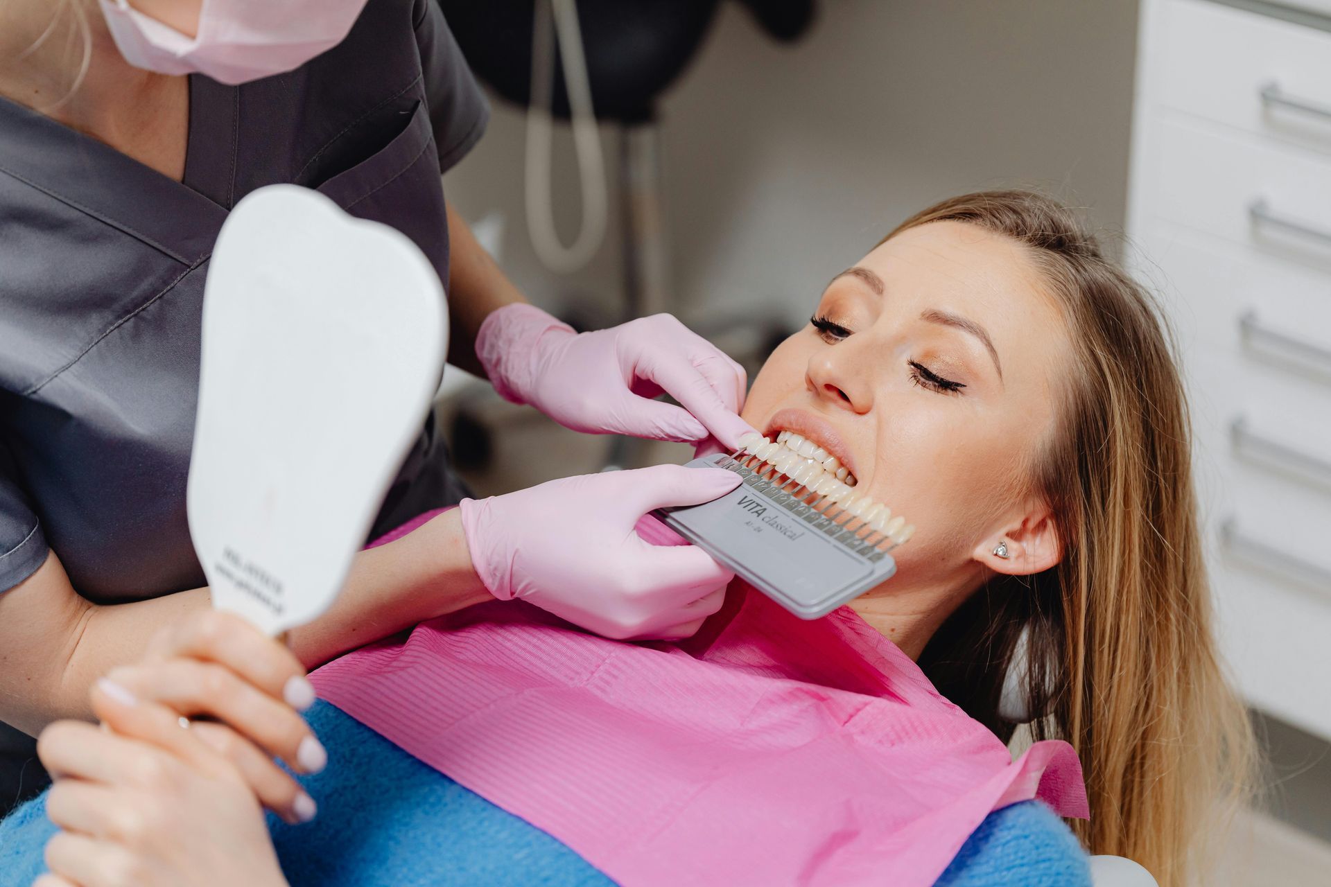 A dental professional holds a color shade guide against a patient's teeth while the patient holds a mirror.