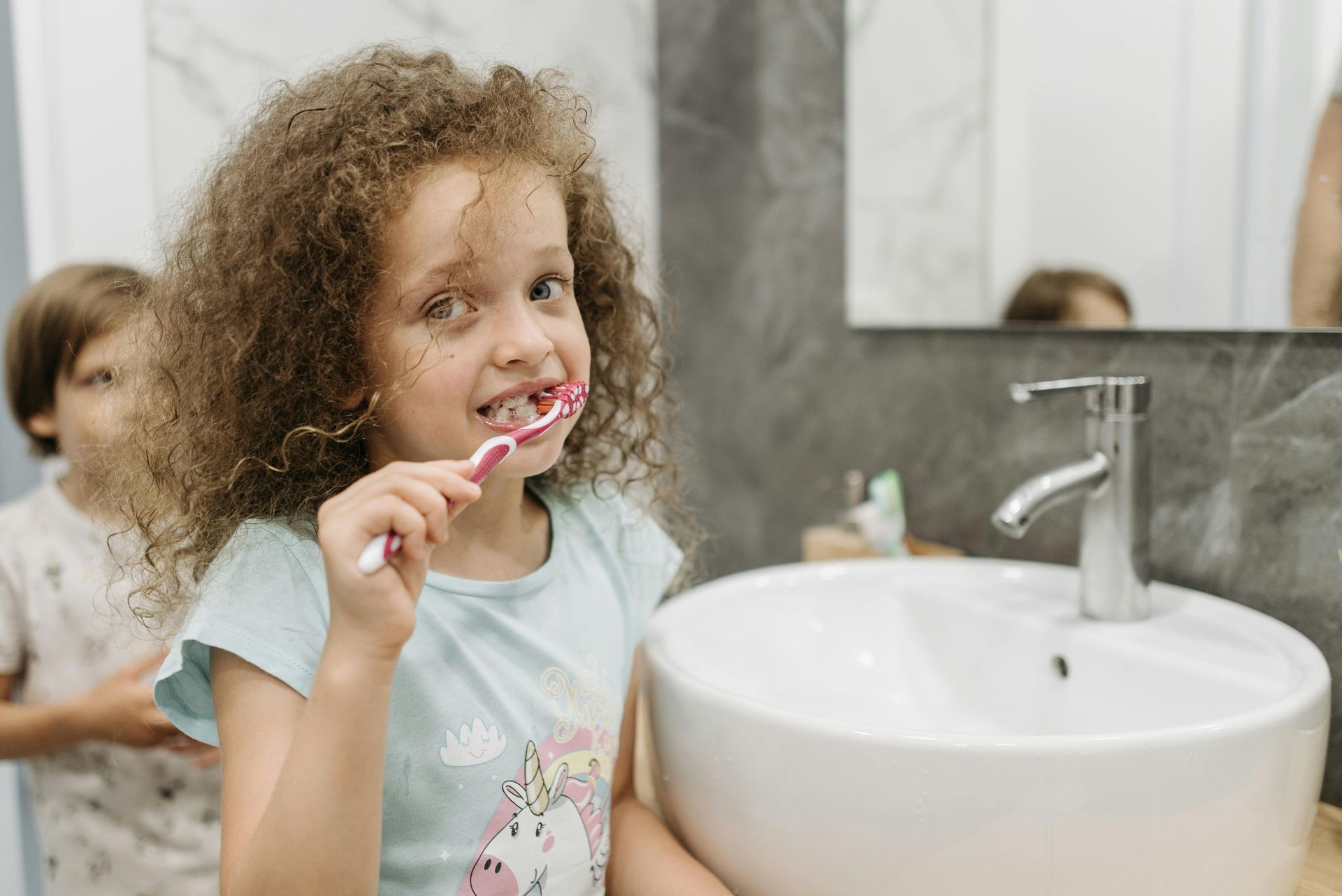 Girl brushing teeth in bathroom, smiling, sink and mirror visible.