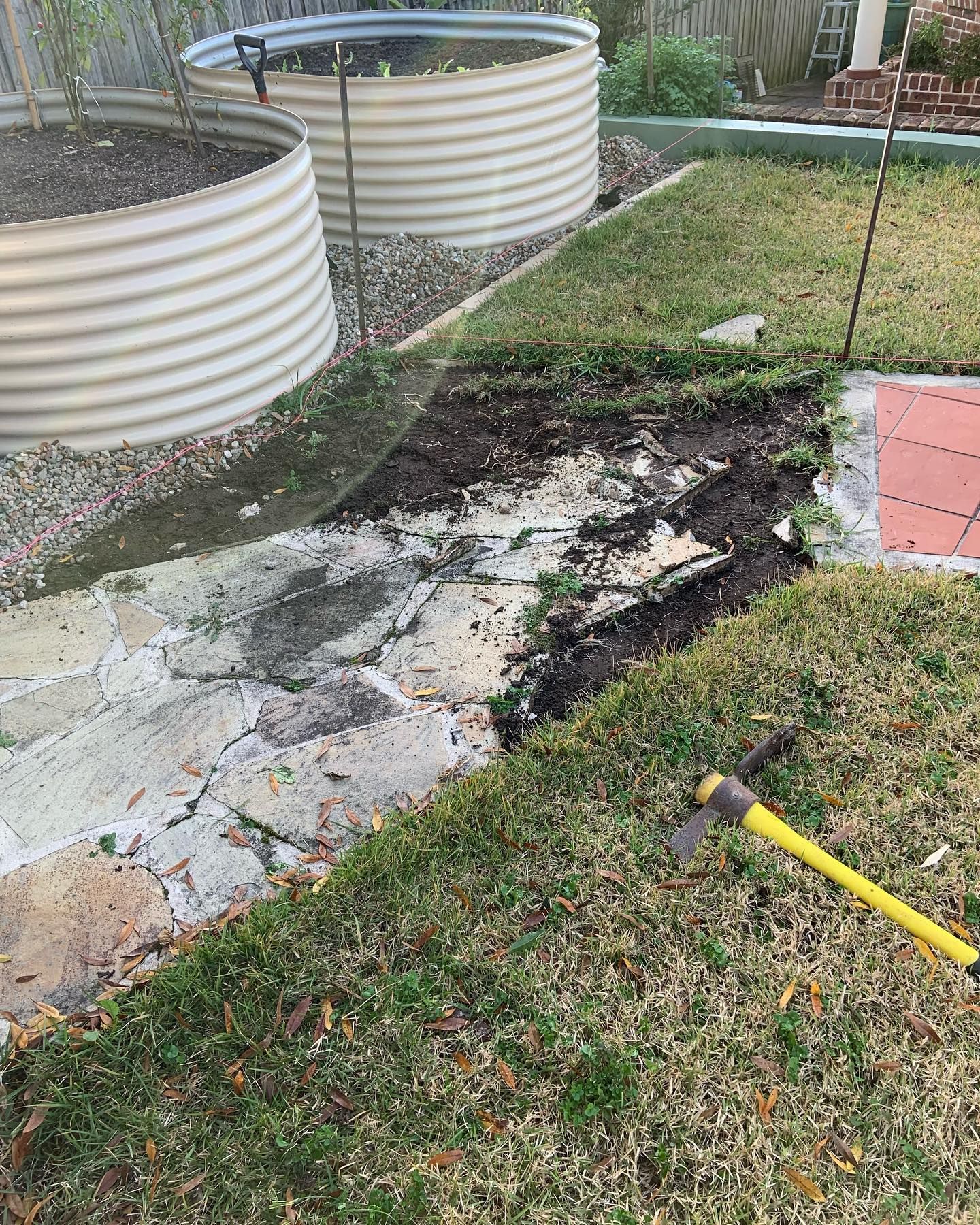 A yellow-handled mattock sits on a lawn beside a flagstone path being cleared of soil and debris near two raised beds.