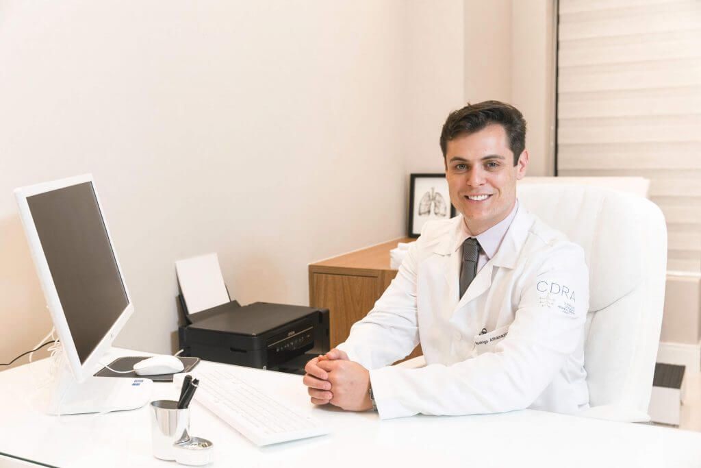 Um homem com jaleco está sentado em uma mesa em frente a um computador.