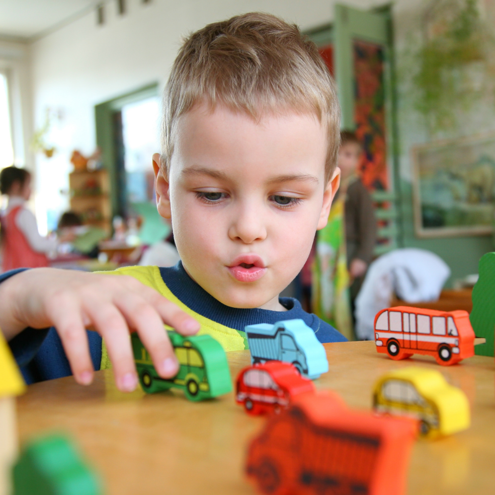 A young boy with light brown hair looks at wooden toy cars on a table in a classroom, his mouth slightly open.