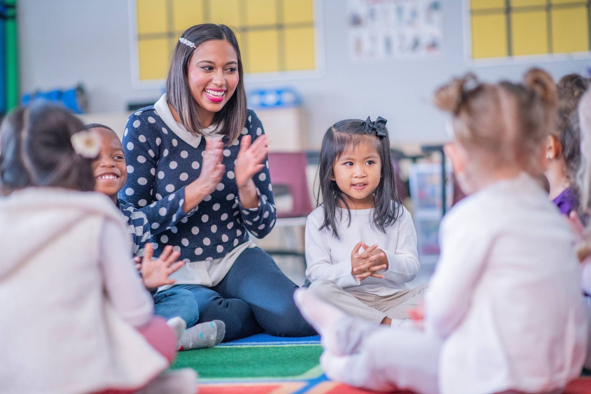 Teacher leading a group of young children in a circle, clapping their hands. They are sitting on a colorful rug in a classroom.