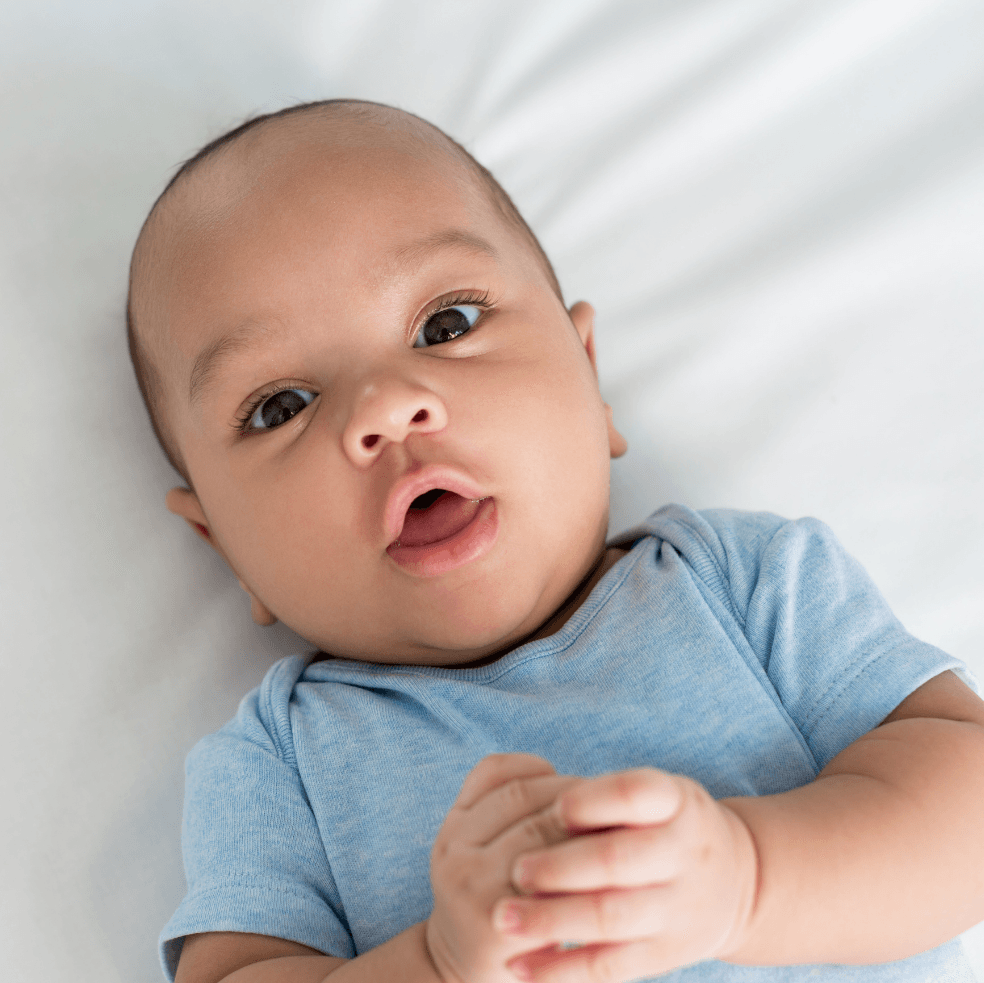 A baby with brown eyes and a blue onesie lies on a white surface with hands clasped.