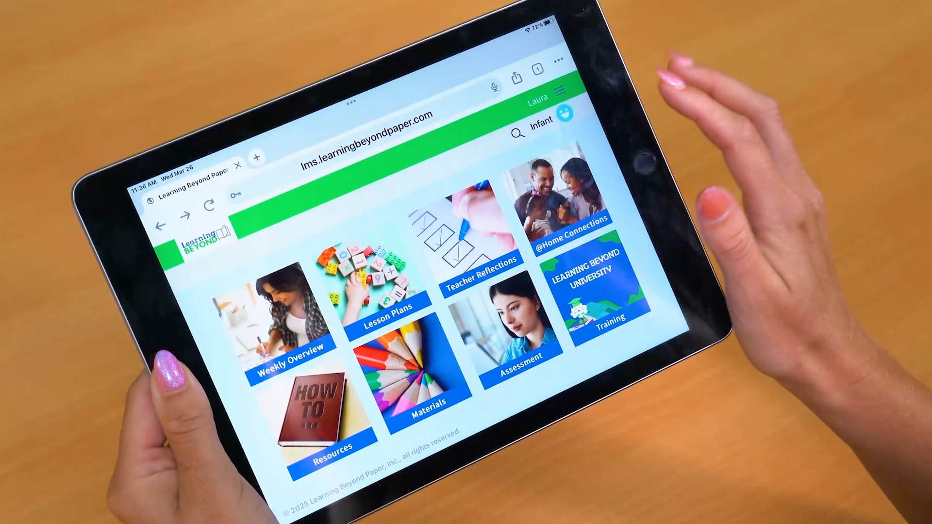 Woman with dark hair, wearing a green vest, smiling while drawing on a tablet at a wooden desk.