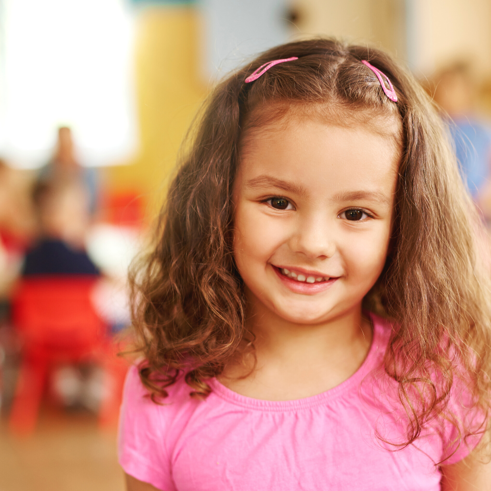 A smiling young girl with brown hair and pink shirt in a classroom setting.